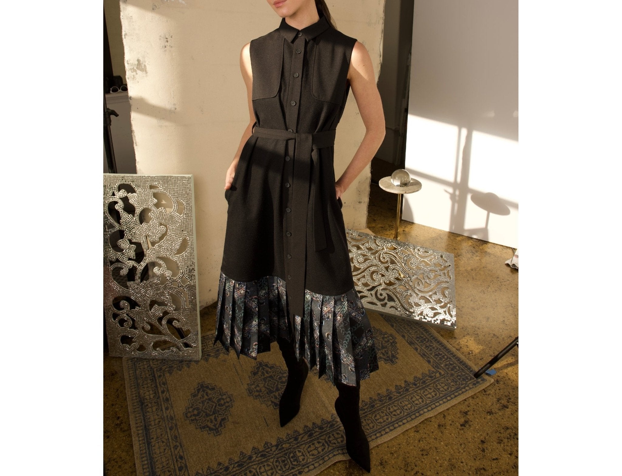 Model wearing a black sleeveless midi shirt dress with tie waist and printed pleated hem, styled with pointed black boots.