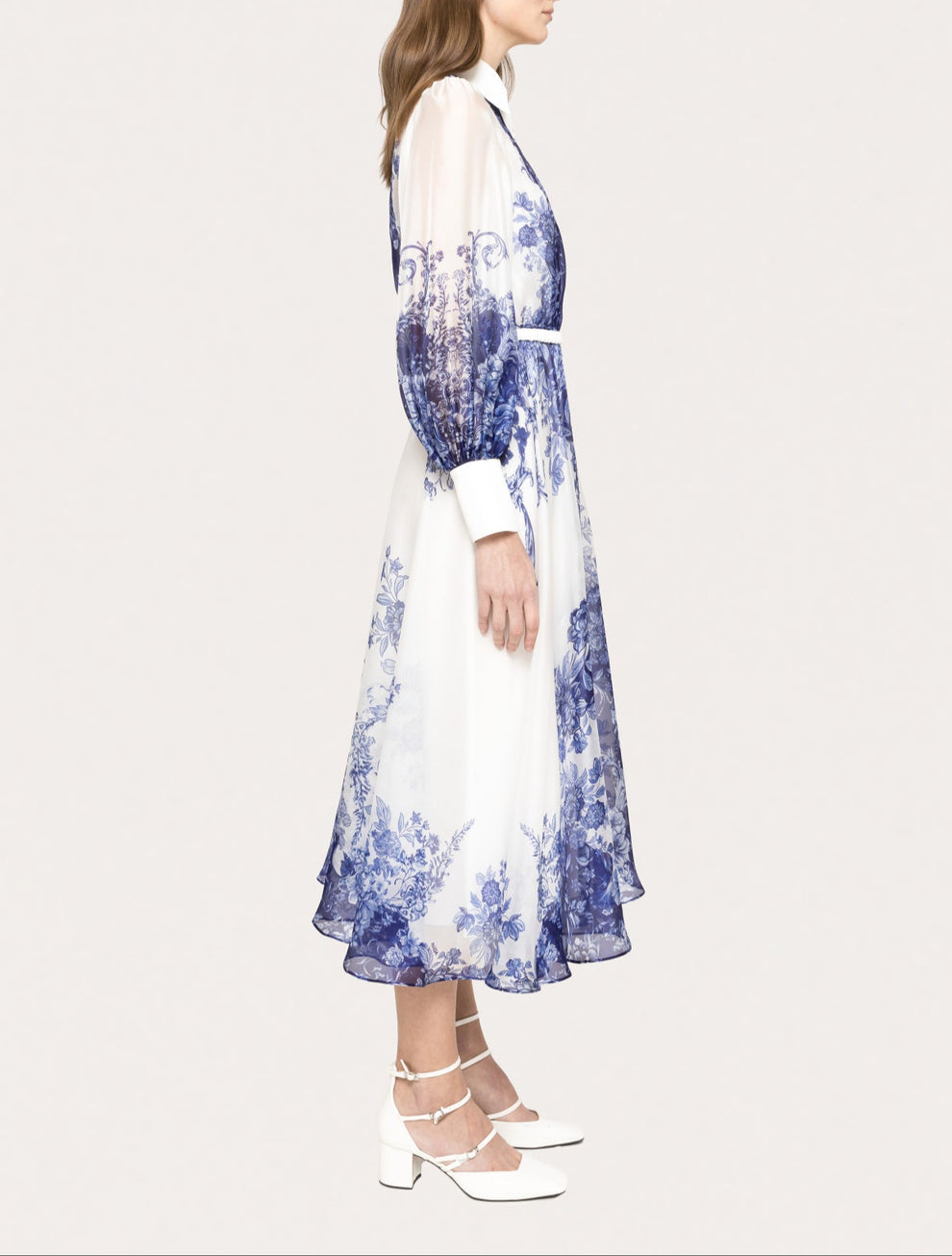 A woman wearing a midi-length blue and white floral print shirt dress with sheer long sleeves, a pointed white collar, button-down front, and a cinched waist, styled with white ankle-strap heels.