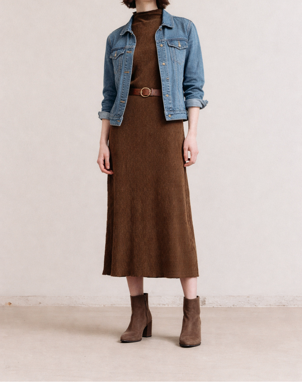 Woman wearing a brown dress, denim jacket, and brown boots against a plain background