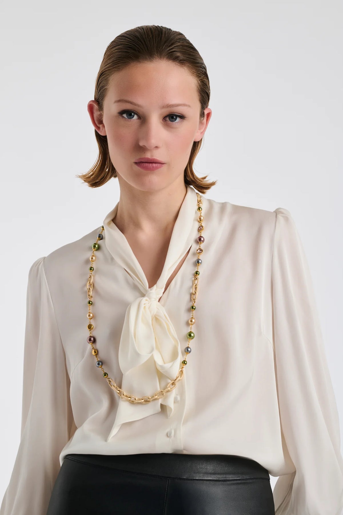 WEILL Paris long gold-tone necklace with green and neutral pearl-look beads, worn as a single strand over an ivory bow blouse.