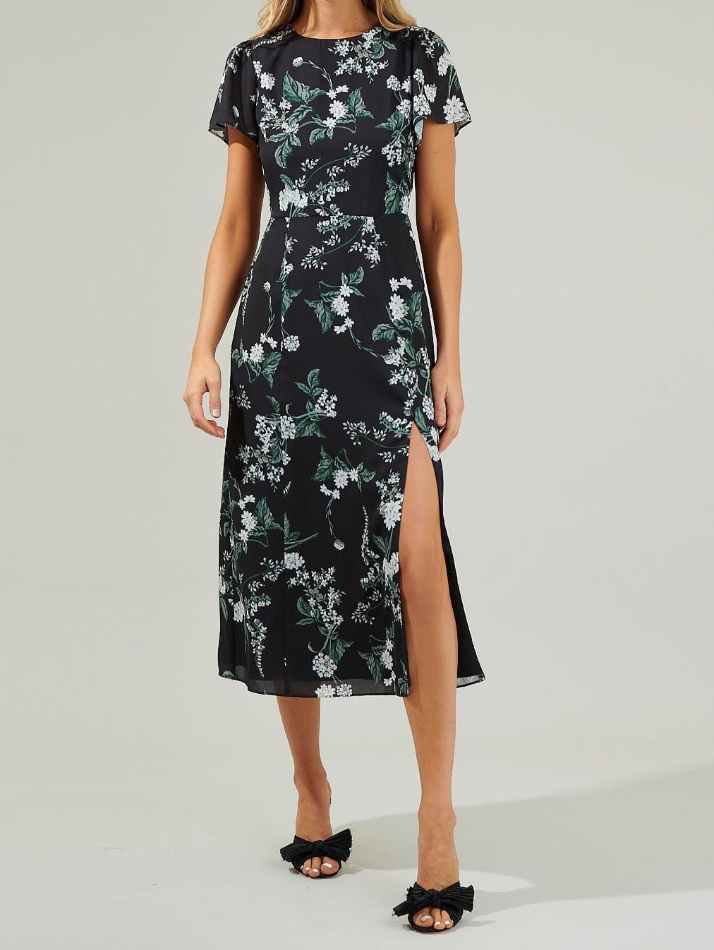 Front view of the Angelonia black floral midi dress with white flowers, green leaves, flutter sleeves and a high side-front slit.