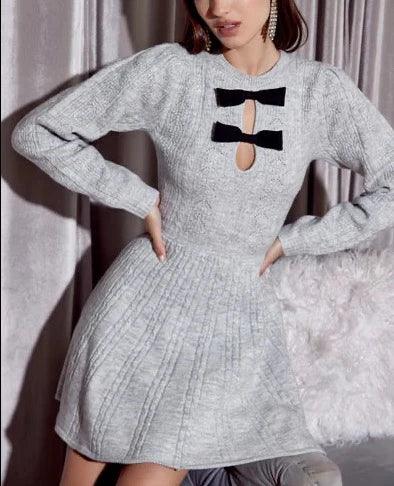 Model wearing the Tessa Bow Front Sweater Knit Dress by Adelyn Rae, featuring a distinctive black bow cut-out detail on the chest, a fitted waist, and a flared skirt, in a soft grey hue, paired with gold earrings.- BTK COLLECTION