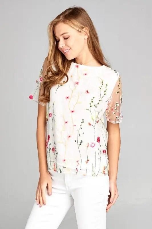 Model wearing a white floral embroidered mesh blouse with sheer short sleeves and a soft lined bodice.