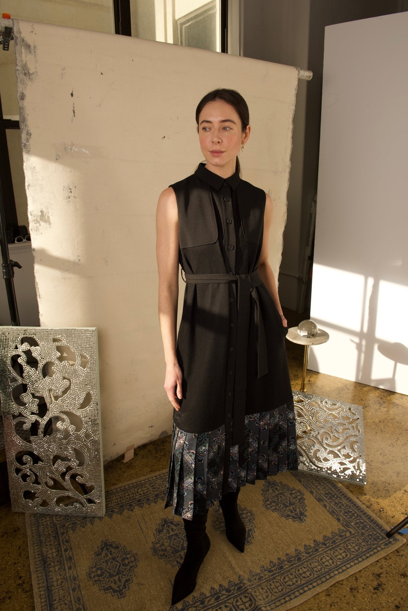 A woman standing in a studio setting, wearing a black Jelda Midi Dress with a collared neckline, button-down front, and a belted waist. The dress features a pleated floral hemline and is accessorized with a black belt and black ankle boots.