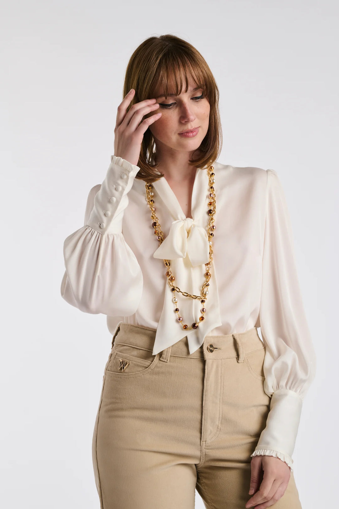 Woman wearing a long double‑chain gold‑tone necklace with brown and pearl‑effect beads over a cream bow‑neck blouse and beige high‑waisted trousers.