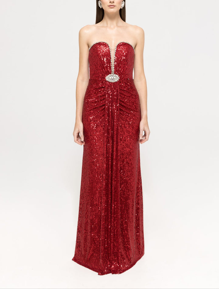 A model is wearing a strapless red sequined maxi dress with a crystal‑embellished plunge neckline and jeweled brooch at the waist, standing against a plain light background.		