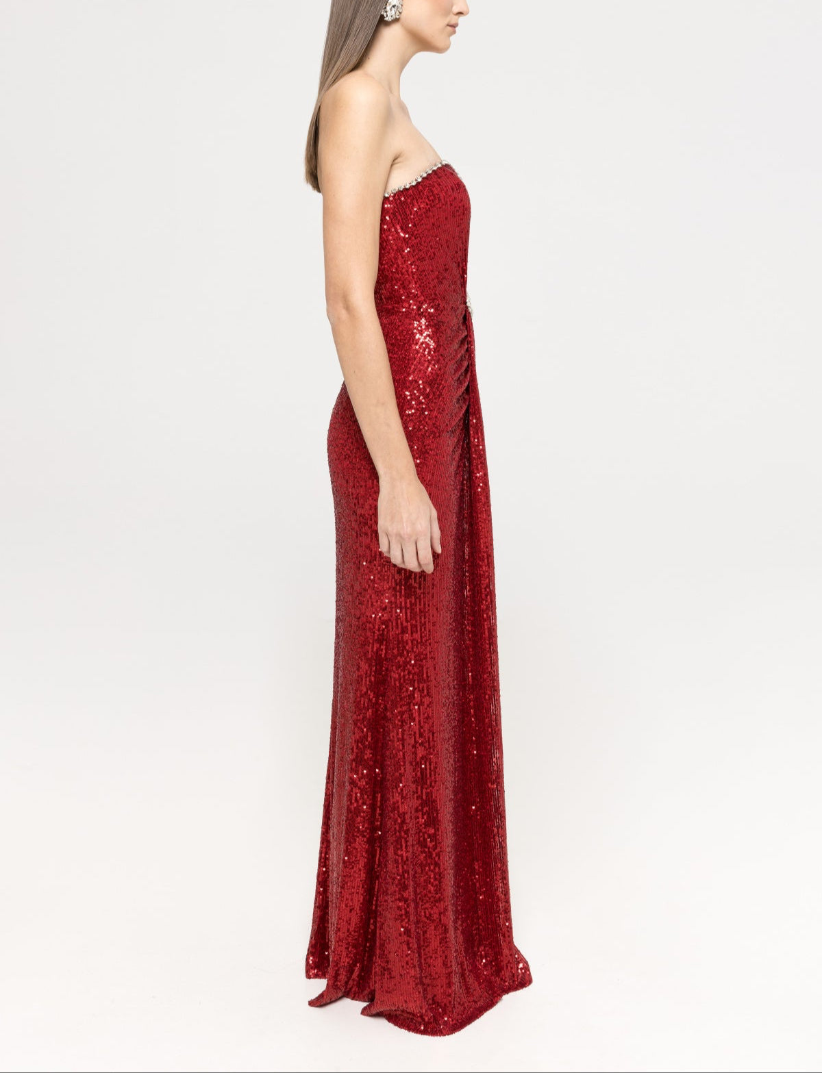 Model is  wearing a strapless red sequin maxi dress with a straight neckline trimmed with crystals and a fitted silhouette, emphasizing the shimmering fabric.