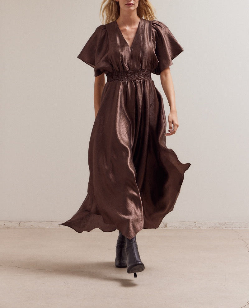 Model walking in flowy unbalanced skirt satin dress and heeled boots in a minimalist setting.