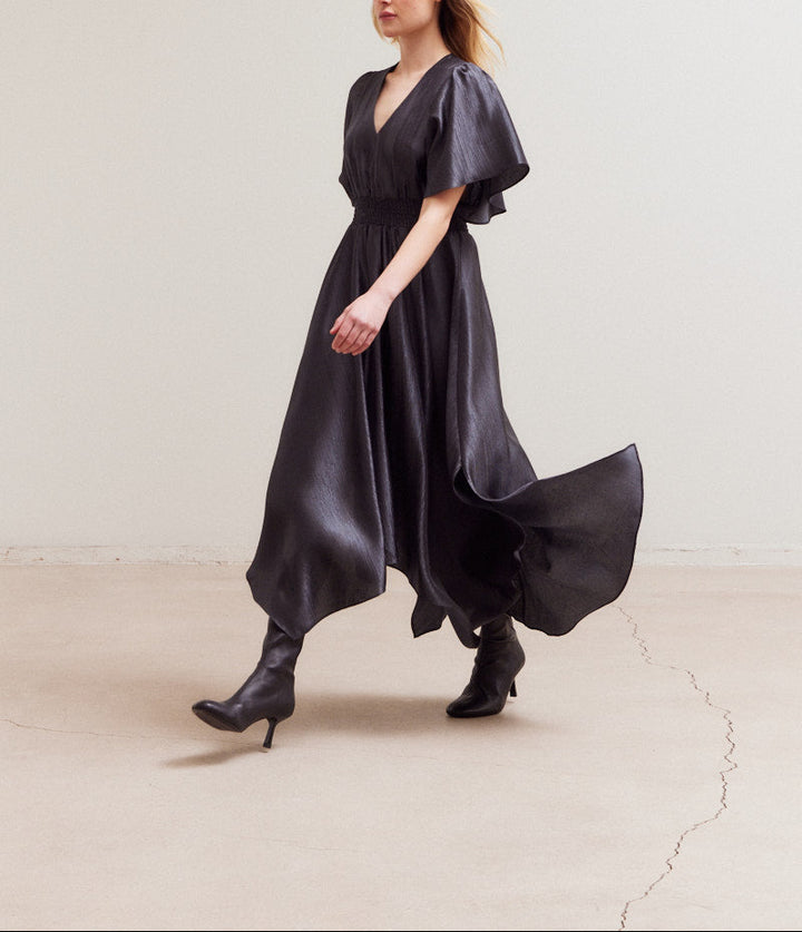 Model walking in flowy handkerchief-hem satin dress and heeled boots in a minimalist setting.