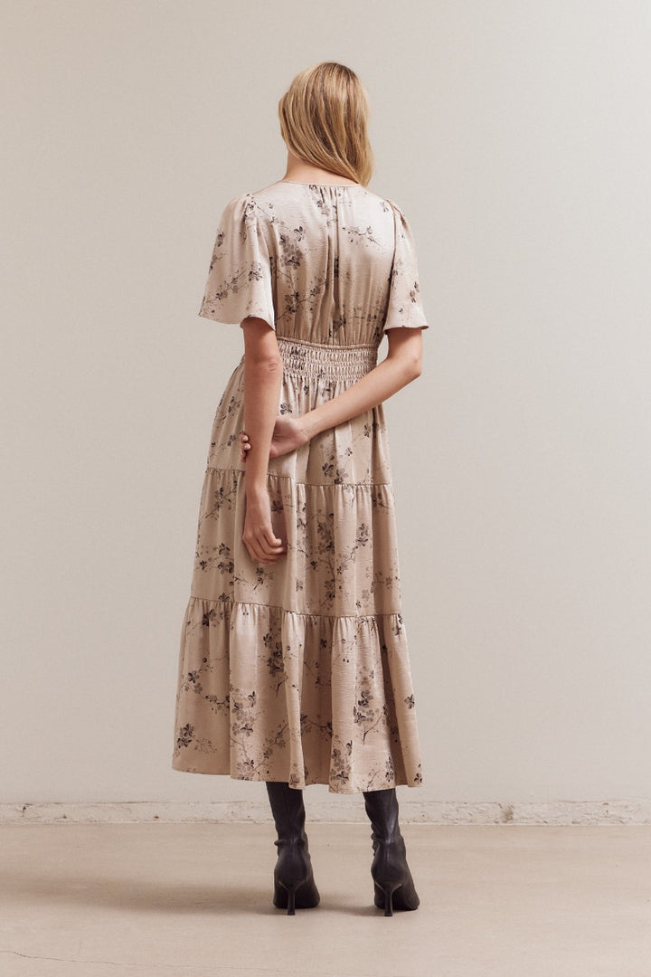 Back view of the natural‑beige floral midi dress with smocked waist and tiered hemline, styled with ankle boots.