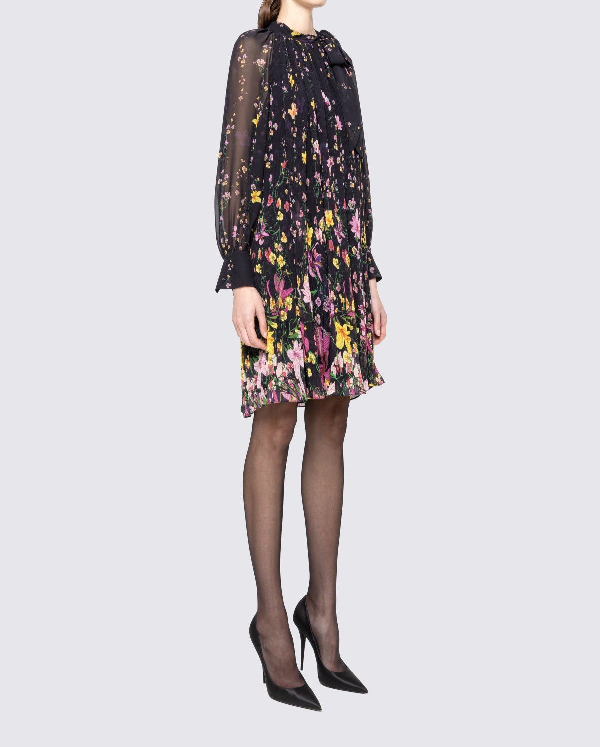 Side view of black floral swing knee length dress featuring bow scarf neck, pleated body and cuffed sheer sleeves