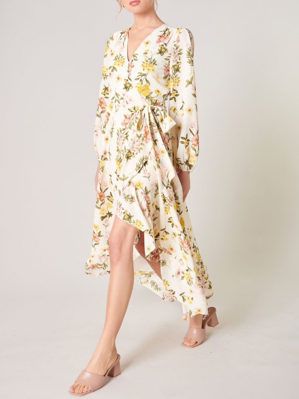 Model wearing the Ashlynn Floral Califa Maxi Wrap Dress by SUGARLIPS, featuring a flowing high-low hemline, long sleeves, and a vibrant print of yellow and pink flowers against a light cream background, complemented by beige strappy heels