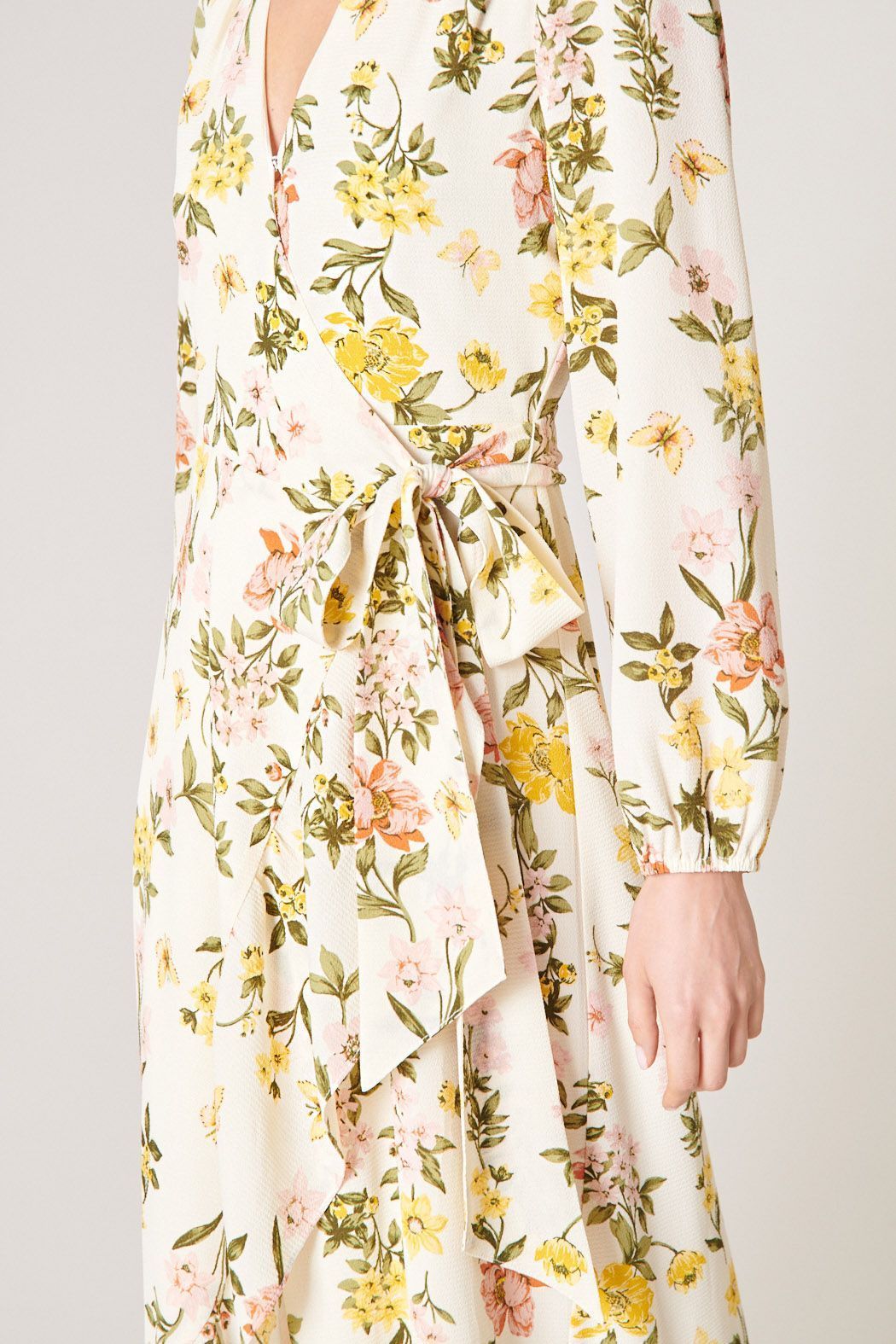 Model wearing the Ashlynn Floral Califa Maxi Wrap Dress by SUGARLIPS, featuring a flowing high-low hemline, long sleeves, and a vibrant print of yellow and pink flowers against a light cream background, complemented by beige strappy heels