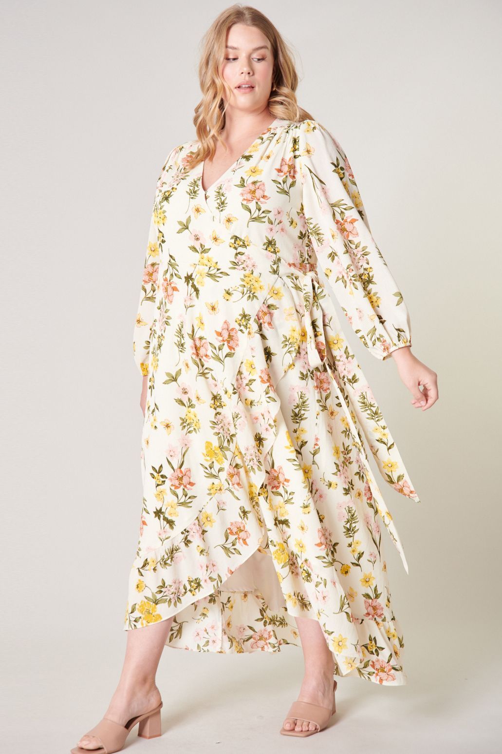Plus Size Model wearing the Ashlynn Floral Califa Maxi Wrap Dress by SUGARLIPS, featuring a flowing high-low hemline, long sleeves, and a vibrant print of yellow and pink flowers against a light cream background, complemented by beige strappy heels
