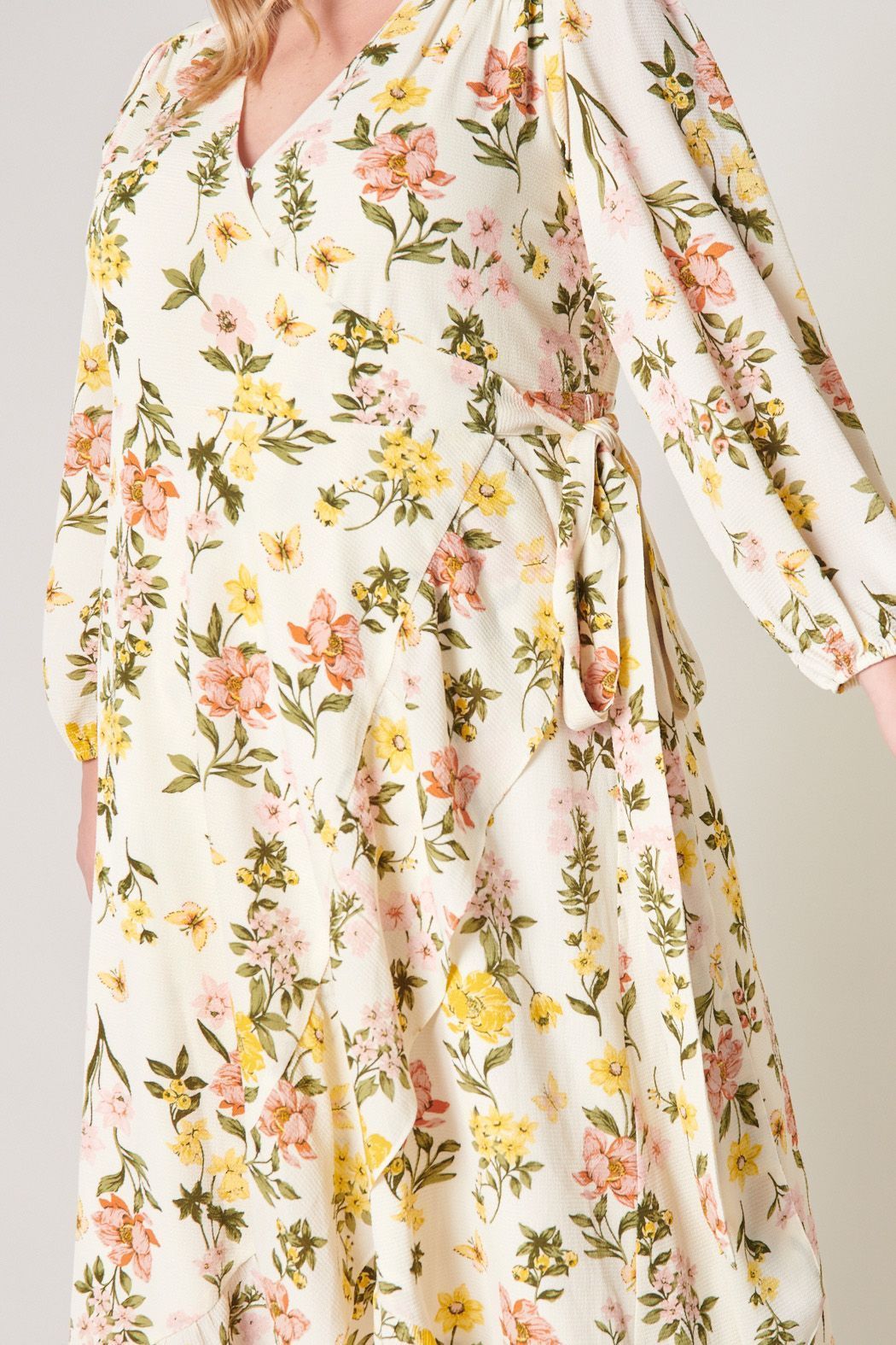 Plus Size Model wearing the Ashlynn Floral Califa Maxi Wrap Dress by SUGARLIPS, featuring a flowing high-low hemline, long sleeves, and a vibrant print of yellow and pink flowers against a light cream background, complemented by beige strappy heels