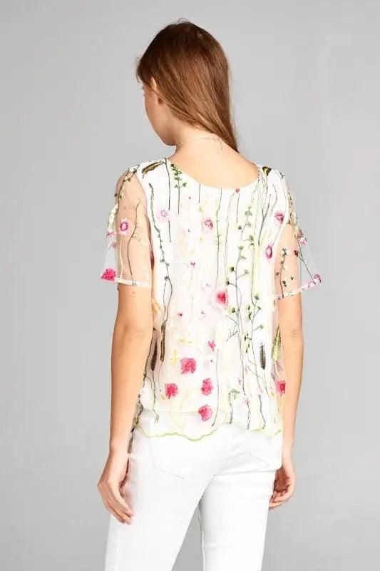 Back view of model wearing a white floral embroidered mesh blouse with sheer short sleeves and a soft lined bodice.